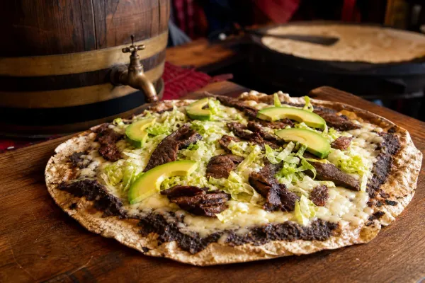 Tlayuda oaxaqueña con asiento ahumado en barril, frijoles refritos, quesillo, tasajo y aguacate